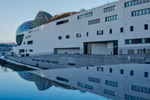 Cette photographie met en scène un bâtiment moderne aux lignes architecturales contemporaines, reflété dans une étendue d’eau calme. Le sujet principal est ce bâtiment, caractérisé par des formes géométriques et des surfaces lisses.La composition joue sur la symétrie et la réflexion, créant une harmonie visuelle. Le cadrage est large, incluant le ciel et les structures environnantes. La lumière, douce et naturelle, semble provenir d’un angle bas, accentuant les contours et les reflets.Les couleurs dominantes sont les tons froids de bleu et gris, renforçant une ambiance minimaliste et épurée. Le style est épuré et architectural, évoquant une ambiance futuriste et sereine.