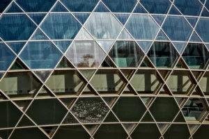 Cette photographie met en scène une façade vitrée géométrique composée de triangles reflétant son environnement. Le sujet principal est la structure architecturale elle-même, avec ses jeux de miroirs et ses lignes anguleuses. La composition repose sur des formes triangulaires répétitives, créant un motif abstrait et dynamique.Le cadrage est serré, mettant en valeur les détails des reflets et des lignes. La lumière est naturelle, probablement en journée, avec des contrastes doux. Les couleurs dominantes sont les teintes bleutées et grises des vitres, ainsi que les reflets verts et blancs de l’environnement extérieur. Le style est épuré et moderne, évoquant une ambiance urbaine et contemporaine.