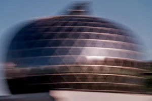 Cette photographie montre un bâtiment aux formes arrondies et horizontales, probablement un dôme ou une structure architecturale moderne. Le sujet principal est flou, suggérant un mouvement ou une vitesse.Le cadrage est serré, mettant en valeur les courbes et les lignes horizontales. La lumière est douce, avec des reflets et des ombres qui créent un effet esthétique. Les couleurs dominantes sont des tons froids et métalliques, avec des nuances grises et bleutées.Le style est dynamique et légèrement abstrait, évoquant une ambiance futuriste et minimaliste.
