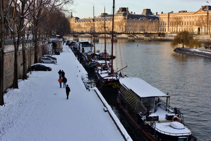 Fabienne · 07/01/2026 · 5488 x 3662 Cette photographie montre un **bassin parisien enneigé**, probablement la Seine. Le **sujet principal** est la **berge couverte de neige**, avec des **bateaux-mouches amarrés** et des **passants**.La **composition** est dynamique, avec une perspective en profondeur, menant vers un **grand bâtiment classique** en arrière-plan. Le **cadrage** est large, capturant à la fois le fleuve et la rive.La **lumière** est douce et hivernale, créant des **tons froids** (blanc, gris). Les **couleurs dominantes** sont le blanc de la neige et les gris bleutés de l’eau et du ciel.L’**ambiance** est calme et poétique, évoquant une **ville endormie sous la neige**. Le style est **réaliste et documentaire**.