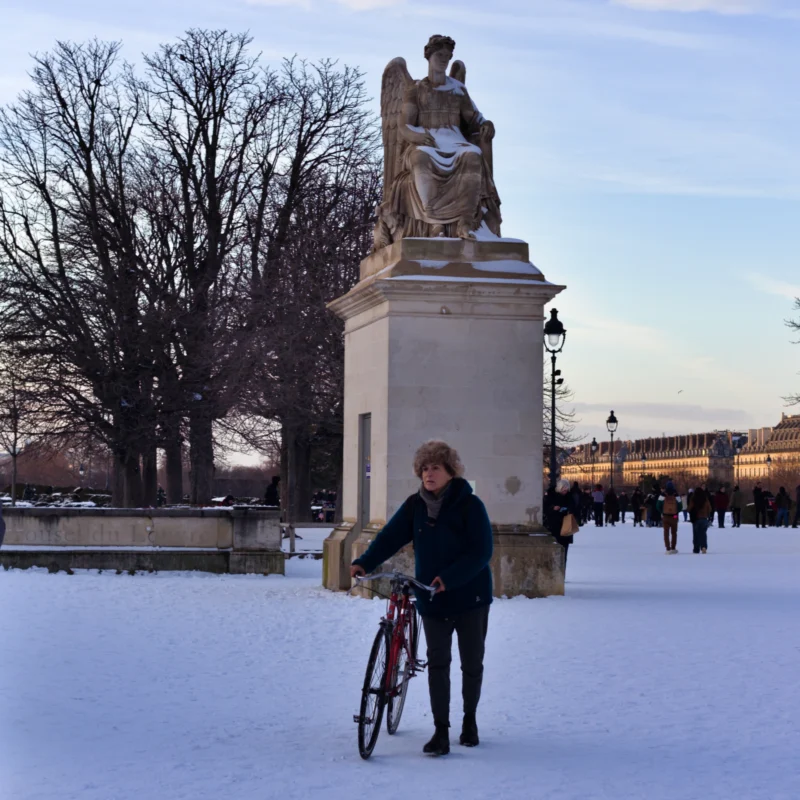 Fabienne · 07/01/2026 · 3273 x 3273 Cette photographie montre une personne tenant un vélo devant une statue monumentale en plein air. Le sujet principal est cette personne, vêtue chaudement, dans un cadre hivernal enneigé.La composition est équilibrée, avec la statue en arrière-plan et la personne en premier plan. Le cadrage est large, incluant des arbres dénudés et des bâtiments à l’horizon. La lumière est douce, typique d’un jour d’hiver, créant des contrastes subtils.Les couleurs dominantes sont les tons froids : blanc de la neige, gris des bâtiments et bleu pâle du ciel. Le style est réaliste et documentaire, évoquant une ambiance calme et paisible, presque mélancolique.