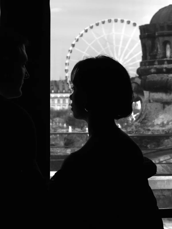 Thomas · 05/01/2026 · 2048 x 2731 Cette photographie en noir et blanc met en scène un **sujet principal flou**, silhouette féminine vue de dos, contemplant une ville. La **composition** oppose cette silhouette à un **Ferris wheel** et des bâtiments emblématiques, suggérant une ambiance urbaine.Le **cadrage** est serré sur la personne, avec une fenêtre encadrant la vue. La **lumière** est douce, créant des contrastes subtils. Les **couleurs dominantes** sont les tons de gris, renforçant une atmosphère mélancolique et nostalgique. Le **style** est poétique et introspectif, évoquant une **ambiance rêveuse et contemplative**.