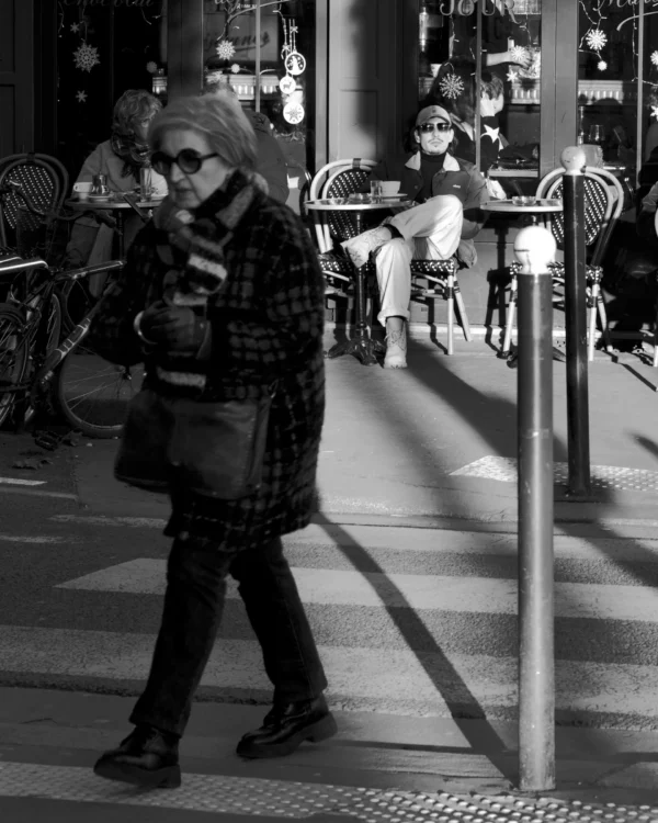 Thomas · 30/12/2025 · 2048 x 2560 Cette photographie en noir et blanc met en scène une personne comme sujet principal, marchant avec un sac à dos et un appareil photo autour du cou. La composition joue sur les contrastes entre ombre et lumière, créant une ambiance dramatique.Le cadrage est serré sur le sujet, avec un arrière-plan flou mais identifiable (bicyclette, personnes assises). La lumière, forte et contrastée, accentue les détails et les textures. Les tons dominants sont les noirs profonds et les gris clairs, typiques du noir et blanc.Le style est documentaire et urbain, évoquant une atmosphère contemplative et mélancolique.