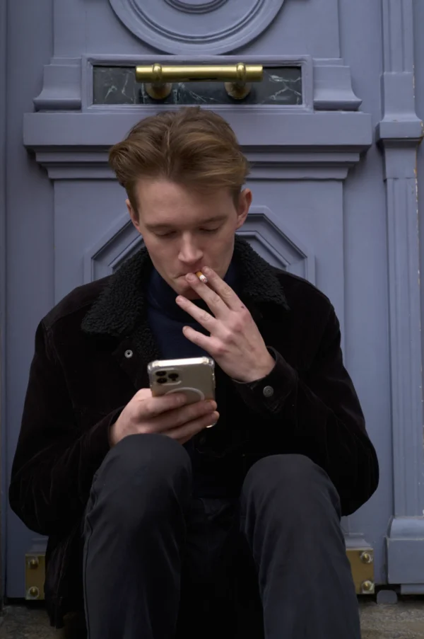 Thomas · 10/01/2026 · 2048 x 3084 Cette photographie montre un jeune homme assis, penché sur son téléphone portable. Le sujet principal est centré, avec un cadrage serré mettant en valeur son visage et ses mains.La composition est simple et naturelle, avec un arrière-plan constitué d'une porte bleue et dorée. La lumière est douce et naturelle, créant des ombres légères sur son visage. Les couleurs dominantes sont les tons froids (bleu, gris) et le noir de son manteau, renforçant une ambiance sobre et moderne. Le style est réaliste et contemporain, évoquant une atmosphère calme et introspective.