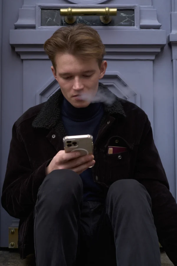 Thomas · 10/01/2026 · 2048 x 3084 Cette photographie montre un jeune homme assis, penché sur son smartphone, dans une posture détendue. Le cadrage est serré, mettant l’accent sur son visage et ses mains.La lumière est douce et naturelle, légèrement voilée, créant une ambiance tamisée. Les couleurs dominantes sont des tons froids et neutres : gris, bleu et noir, renforçant une atmosphère sobre et moderne. Le style est épuré et contemporain, évoquant une scène urbaine et intime. L’ambiance est calme et légèrement mélancolique, reflétant une solitude moderne.