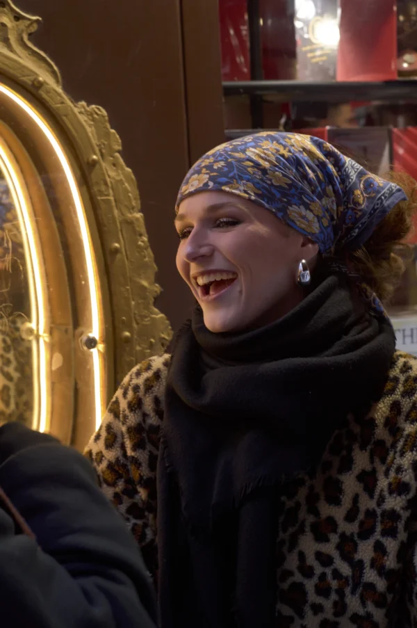 Thomas · 10/01/2026 · 2048 x 3084 Cette photographie met en scène une femme comme sujet principal, souriante et vêtue d’un turban coloré et d’une veste en fourrure synthétique. Le cadrage est serré, centré sur son visage et sa poitrine.La lumière semble douce et naturelle, éclairant son visage avec des reflets chauds. Les couleurs dominantes sont les tons froids du turban (bleus, violets) contrastant avec les teintes chaudes de la fourrure et des éléments dorés en arrière-plan.L’ambiance est chaleureuse et festive, évoquant une scène conviviale, peut-être lors d’un événement hivernal ou culturel.