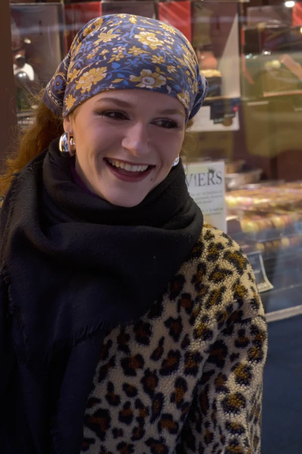 Thomas · 10/01/2026 · 2048 x 3084 Cette photographie met en scène une jeune femme souriante, sujet principal, vêtue d’un manteau à motifs léopard et d’un foulard coloré. Le cadrage est serré, centré sur elle, avec un arrière-plan flou suggérant un marché ou une boutique.La lumière semble naturelle et douce, éclairant son visage. Les couleurs dominantes sont vives : les teintes chaudes du foulard (bleus, jaunes) contrastent avec les tons neutres du manteau. L’ambiance est chaleureuse et conviviale, évoquant une scène de vie quotidienne joyeuse et colorée.
