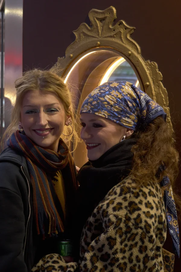 Thomas · 10/01/2026 · 2048 x 3084 Cette photographie met en scène deux femmes souriantes en ambiance festive nocturne. Le sujet principal est la femme à droite, déguisée en lionne avec une crinière en fourrure et un masque doré, tandis que l’autre l’observe avec enthousiasme.La composition est serrée, centrée sur elles. Le cadrage est rapproché, créant une intimité. La lumière est douce, légèrement tamisée, avec des éclairages artificiels ambiants. Les couleurs dominantes sont les tons chauds (or, marron) et les contrastes vifs (fourrure noire, bleu du masque).L’ambiance est joyeuse et festive, évoquant une soirée déguisée ou un événement nocturne.