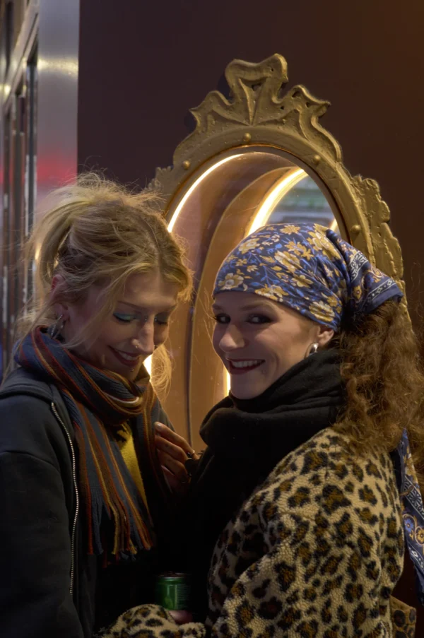 Thomas · 10/01/2026 · 2048 x 3084 Cette photographie met en scène deux femmes comme sujets principaux. Elles sont cadrées en gros plan, l’une souriante avec un chapeau orné et un foulard coloré, l’autre portant un manteau à motifs léopard.La lumière semble douce et naturelle, éclairant leurs visages. Les couleurs dominantes sont les tons chauds et vifs : bleu, orange, noir et motifs graphiques. Le style est spontané et naturel, évoquant une ambiance conviviale et festive, probablement lors d’un événement nocturne ou une fête.