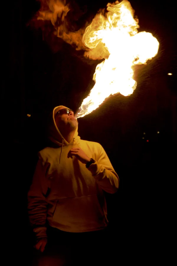 Thomas · 24/01/2026 · 2048 x 3080 Cette photographie met en scène un homme comme sujet principal, manipulant une torche à feu produisant une flamme orange et jaune vive. Le cadrage est serré, centré sur son visage et ses mains, isolant ainsi l'action.La lumière est dramatique, avec un éclairage contrasté où la flamme domine, créant une ambiance intense et mystérieuse. Les couleurs dominantes sont les tons chauds (oranges, jaunes) de la flamme, contrastant avec les teintes sombres de l'arrière-plan.Le style est réaliste et spontané, évoquant une scène captée sur le vif, renforçant une ambiance à la fois spectaculaire et légèrement inquiétante.