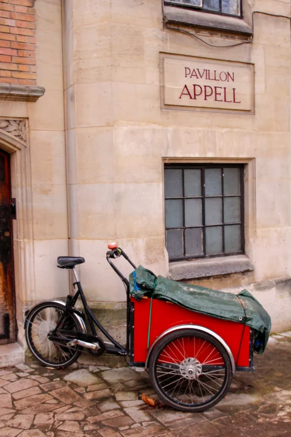 Cédric · 17/01/2026 · 1365 x 2048 Cette photographie met en scène un vélo-cargo rouge et vert, sujet principal, garé devant un bâtiment ancien. La composition est simple et équilibrée, avec le vélo centré et légèrement de biais.Le cadrage est serré, mettant en valeur les détails du vélo et de la façade. La lumière douce, probablement naturelle, adoucit les contours et crée des ombres douces. Les couleurs dominantes sont les tons rouges, verts et gris, renforçant une ambiance chaleureuse et vintage.Le style évoque un charme rétro et artisanal, tandis que l’ambiance est paisible et intemporelle, évoquant un lieu historique et apaisant.