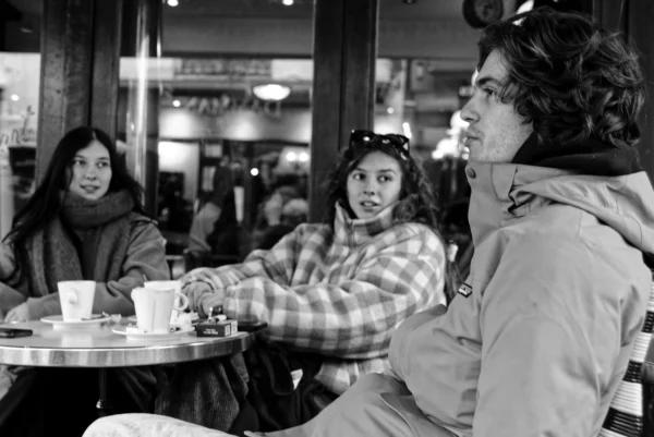 Mickaël · 10/01/2026 · 4530 x 3024 Cette photographie en noir et blanc capture un moment intime dans un café. Le sujet principal est un trio assis autour d'une table : une femme souriante au centre, entourée d’une personne à gauche et d’un homme à droite, tous engagés dans une conversation.La composition est serrée, centrée sur les visages et les mains, créant une ambiance conviviale. Le cadrage est légèrement rapproché, mettant l’accent sur les expressions et les détails des vêtements. La lumière douce, probablement naturelle filtrée par les vitres, adoucit les contours et ajoute une touche chaleureuse.L’absence de couleurs renforce l’aspect nostalgique et universel de la scène, évoquant une ambiance détendue et intime.