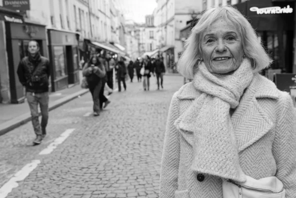 Mickaël · 10/01/2026 · 5336 x 3562 Cette photographie en noir et blanc met en scène une femme âgée au premier plan, vêtue d’un long manteau et d’un écharpe, souriante et regardant l’appareil. La composition joue sur le contraste entre elle et les passants floutés en arrière-plan, créant une profondeur de champ.Le cadrage serré sur son visage et la lumière douce, probablement naturelle, accentuent son expression. Les tons clairs et ombres renforcent l’ambiance calme et mélancolique. Le style est documentaire, capturant un moment de vie urbaine authentique, avec une touche d’émotion et de nostalgie.