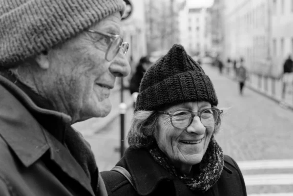 Mickaël · 10/01/2026 · 4180 x 2790 Cette photographie en noir et blanc met en scène deux personnes âgées comme sujets principaux, souriantes et en conversation. La composition est serrée, centrée sur leurs visages, avec un cadrage rapproché. L’arrière-plan flou montre une rue légèrement animée.La lumière est douce et naturelle, sans contraste marqué. Les tons dominants sont les gris et noirs, typiques du noir et blanc, accentuant les textures des vêtements et des visages.Le style est documentaire et intimiste, évoquant une ambiance chaleureuse et authentique, pleine d’humanité et de simplicité.