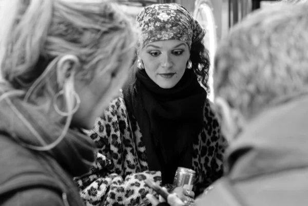 Mickaël · 10/01/2026 · 5023 x 3353 Cette photographie en noir et blanc met en scène une femme au centre, vêtue d’un costume élégant et coiffée d’un chapeau orné, comme une starlette ou une actrice. Elle semble être l’attraction principale, entourée de personnes attentives.La composition est serrée, avec un cadrage en plan moyen, mettant l’accent sur les interactions et les expressions. La lumière est douce, créant des contrastes subtils et des ombres légères. L’ambiance est intimiste et chaleureuse, évoquant une scène de rencontre ou de célébration. Le style rappelle le cinéma classique ou un événement mondain.