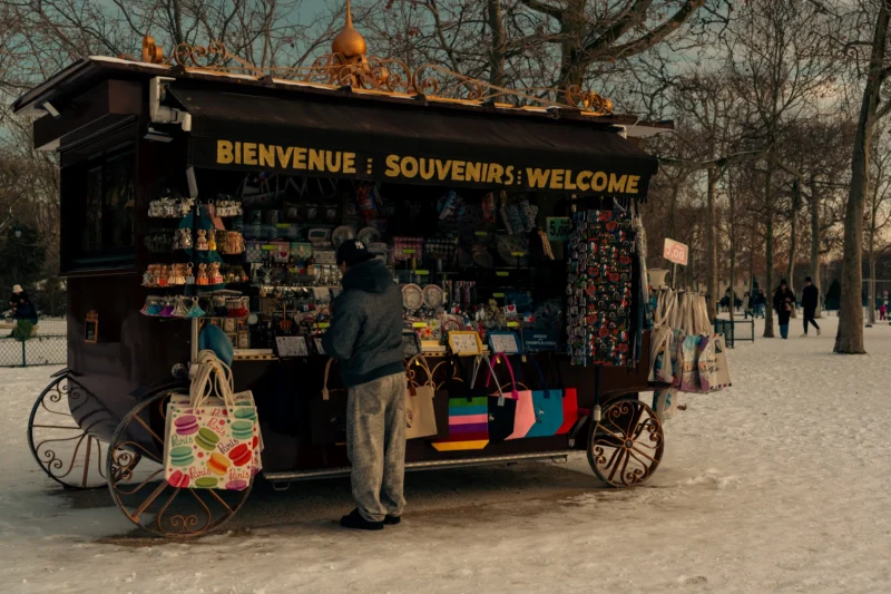 Don · 6706 x 4471 Cette photographie montre un stand de souvenirs itinérant dans un parc. Le sujet principal est un vendeur, dos à la caméra, devant son étalage rempli d’objets colorés.La composition est symétrique, avec le stand central et des passants en arrière-plan. Le cadrage est serré, mettant en valeur les détails du stand et son ambiance chaleureuse. La lumière est naturelle et douce, typique d’un jour ensoleillé d’hiver.Les couleurs dominantes sont les tons froids (gris du sol) contrastant avec les couleurs vives des souvenirs. Le style est réaliste et documentaire, évoquant une ambiance authentique et conviviale.