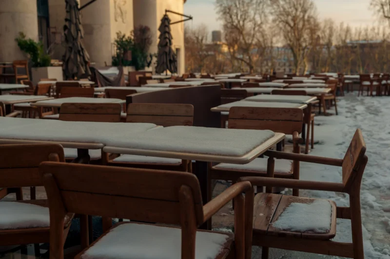 Don · 6695 x 4463 Cette photographie montre une série de tables et chaises en bois disposées à l'extérieur, probablement sur une terrasse. Le sujet principal est l’espace vide et ordonné des tables recouvertes de nappes blanches.La composition est symétrique et répétitive, avec un cadrage large incluant un bâtiment en arrière-plan. La lumière, douce et tamisée, suggère un moment crépusculaire ou hivernal. Les couleurs dominantes sont les tons froids : gris, blanc et bois naturel.Le style est réaliste et sobre, évoquant une ambiance calme et presque mélancolique, renforcée par la neige légère et l’absence de vie.