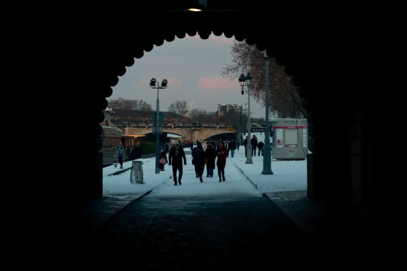 Don · 5322 x 3548 Cette photographie capture un groupe de personnes traversant un passage arché sous un arc sombre. Le sujet principal est l’activité humaine en mouvement, vue à travers un cadrage en tunnel qui isole la scène.La composition utilise un jeu d’ombres et de lumière naturelle, avec des teintes chaudes (orange, rose) en arrière-plan, contrastant avec les tons froids et sombres de l’avant-plan. Les couleurs dominantes sont les bleus grisâtres et les reflets dorés du coucher de soleil.Le style est réaliste et légèrement cinématographique, créant une ambiance mélancolique et contemplative, presque onirique.