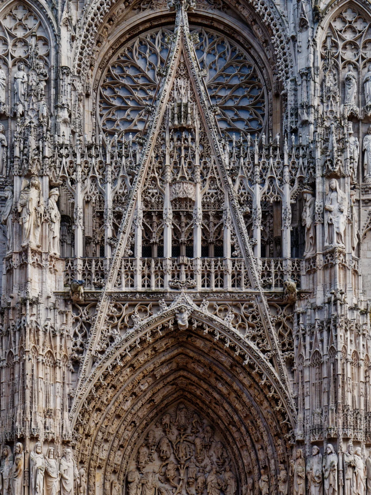 Daniel · 17/03/2022 · 2533 x 3377 Cette photographie met en scène une façade de cathédrale gothique. Le sujet principal est un portail richement sculpté, surmonté d'une rosace et de vitraux colorés.La composition est verticale, centrée sur l’architecture complexe, avec des arcs brisés et des détails ornés. Le cadrage serré met en valeur les éléments architecturaux.La lumière naturelle éclaire les sculptures, créant des contrastes entre ombres et lumières. Les couleurs dominantes sont les tons gris-beige des pierres, rehaussés par les teintes bleutées et rouges des vitraux.Le style est gothique, évoquant une ambiance solennelle et majestueuse.