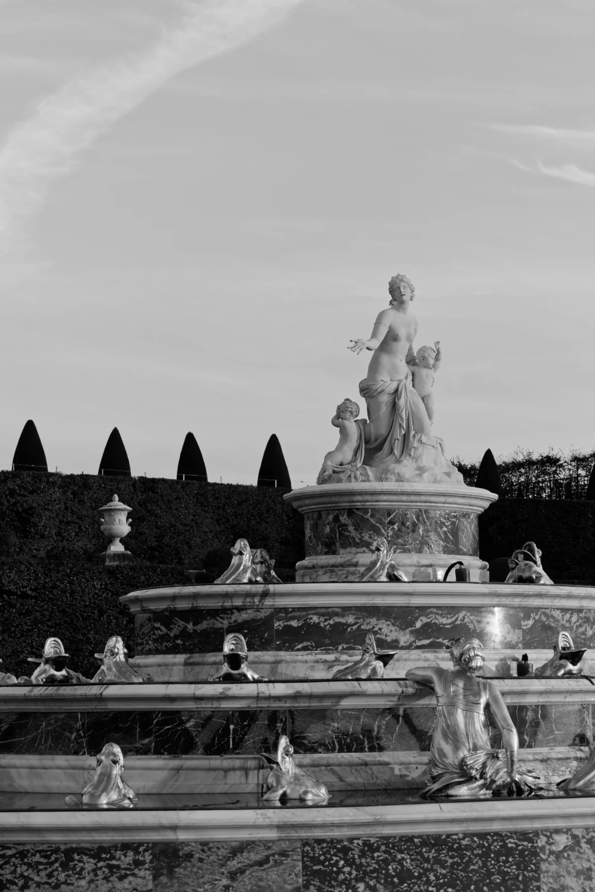 Daniel · 12/10/2015 · 2765 x 4144 Cette photographie en noir et blanc met en scène une fontaine monumentale comme sujet principal, avec une statue centrale représentant une figure allégorique entourée de personnages. La composition est symétrique et hiérarchisée, avec plusieurs niveaux de bassins et de sculptures.Le cadrage est serré sur la fontaine, intégrant partiellement des éléments végétaux en arrière-plan. La lumière douce, probablement naturelle, crée des contrastes subtils et adoucit les contours. Les tons dominants sont les gris, renforçant une ambiance classique et intemporelle. Le style est artistique et élégant, évoquant une atmosphère paisible et majestueuse.