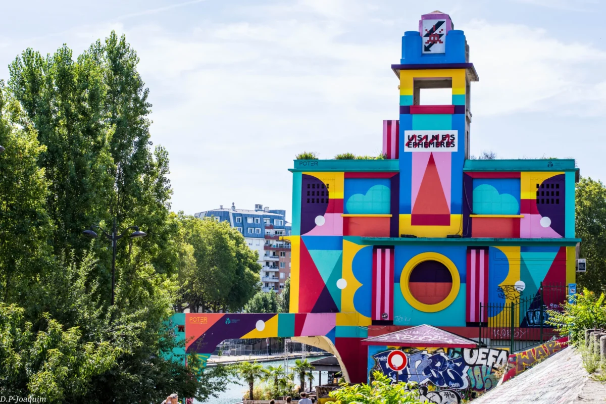 Pierre-Joaquim · 30/03/2015 · 6000 x 4000 Cette photographie montre un bâtiment coloré et ludique, **la Tour des Dames** à Paris, située dans le Jardin Vélizy. Le sujet principal est cette structure en forme de puzzle, recouverte de motifs géométriques et de couleurs vives.La composition met en valeur la tour au centre, entourée de végétation. Le cadrage inclut une partie des arbres et du ciel, créant un contraste entre nature et architecture. La lumière est naturelle, éclatante, mettant en relief les teintes saturées. Les couleurs dominantes sont le **rose, bleu, jaune et vert**, dynamiques et joyeuses. Le style est **artistique et enfantin**, évoquant une ambiance festive et créative.