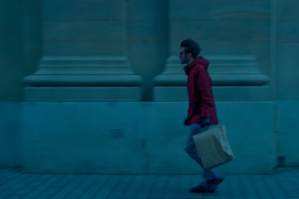 Don · 5558 x 3705 Cette photographie met en scène une personne en mouvement, vêtue d’un manteau rouge, tenant une valise. Le sujet principal est légèrement flou, suggérant un cadrage dynamique.La composition est épurée, avec un premier plan sombre et des colonnes architecturales en arrière-plan. Le cadrage est serré, centré sur le personnage.Les couleurs dominantes sont les teintes froides (bleu dominant) et le rouge vif du manteau. La lumière est tamisée, créant une ambiance mystérieuse et légèrement mélancolique, typique d’un style cinématographique ou artistique moderne.