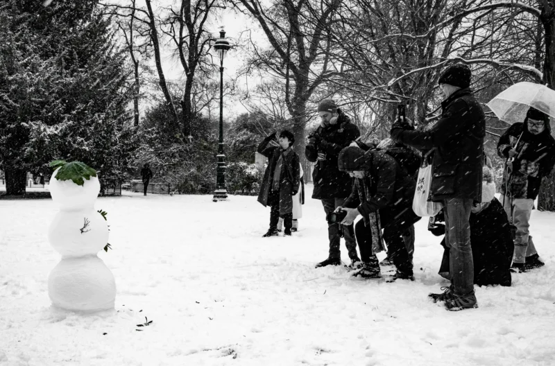 Taunia · 2500 x 1650 Cette photographie en noir et blanc capture un groupe de personnes en hiver dans un parc enneigé. Le **sujet principal** est un **bonhomme de neige** à gauche, tandis que les personnes, vêtues chaudement, semblent discuter ou observer.La **composition** est équilibrée, avec un **cadrage large** incluant un chemin et des arbres dénudés. La **lumière** est douce, probablement naturelle et diffuse, créant des contrastes subtils. Les **couleurs dominantes** sont les nuances de gris, renforçant l’ambiance hivernale.Le **style** est documentaire et sobre, évoquant une **ambiance calme et nostalgique**, typique d’un jour froid et paisible.