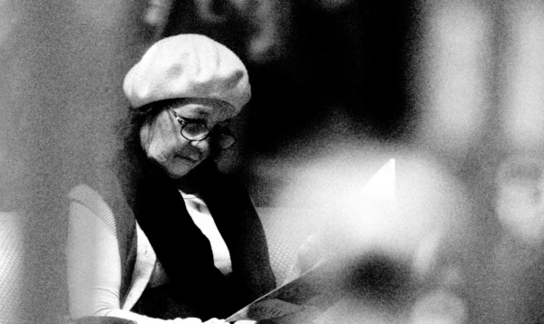 Franck T. · 24/01/2026 · 1400 x 836 Cette photographie en noir et blanc montre une personne, probablement Yoko Ono, comme sujet principal. Elle porte un chapeau et une écharpe, tenant un micro ou un objet similaire. Le cadrage est serré, mettant l’accent sur son visage et ses mains.La lumière est douce, créant des contrastes modérés. L’ambiance est intime et artistique, évoquant une scène musicale ou une performance. Les tons dominants sont les gris et noirs, renforçant l’aspect rétro et contemplatif. Le style rappelle les photographies des années 1960-1970, avec une touche d’authenticité et de simplicité.