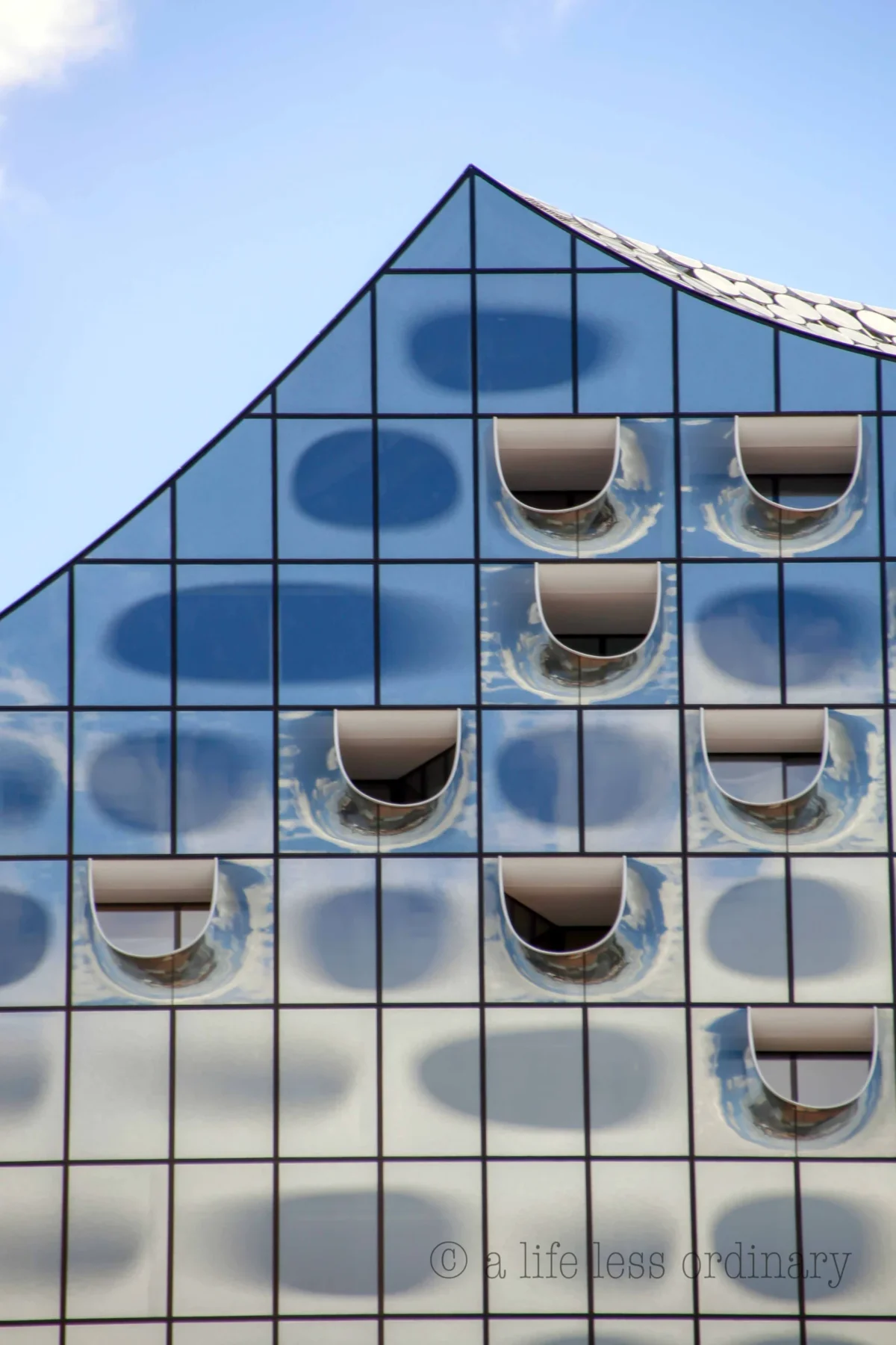 Cédric · 17/06/2017 · 3362 x 5043 Cette photographie met en scène une façade vitrée en forme de triangle, reflétant des objets circulaires sombres, probablement des casques ou des éléments sphériques. La composition repose sur une symétrie géométrique et une répétition harmonieuse des motifs.Le cadrage est serré, centré sur la structure, avec un angle légèrement bas. La lumière est naturelle, vive et contrastée, accentuant les reflets. Les couleurs dominantes sont les tons bleus du ciel et les teintes grises des vitres et des objets reflétés.Le style est épuré et moderne, évoquant une ambiance architecturale contemporaine et minimaliste.