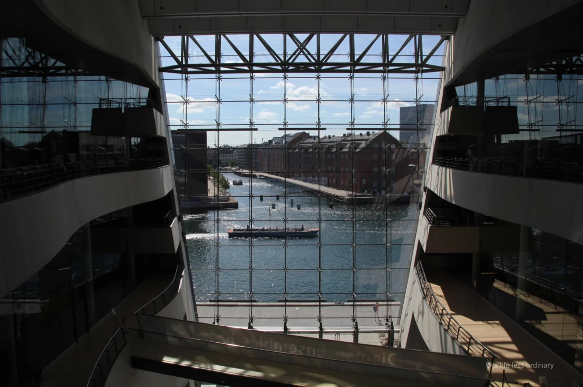 Cédric · 09/08/2013 · 5067 x 3366 Cette photographie montre un bâtiment moderne avec une vue intérieure depuis un escalier en colimaçon. Le sujet principal est un bassin d’eau entouré de structures architecturales en verre et béton, reflétant une ambiance urbaine.La composition est symétrique et dynamique, avec des lignes droites et des angles géométriques. Le cadrage est large, incluant des balustrades et des espaces vides. La lumière naturelle, filtrée par les grandes baies vitrées, crée des contrastes nets entre ombres et clarté. Les couleurs dominantes sont les tons gris (béton, verre) et bleus (eau, ciel).Le style est épuré et contemporain, évoquant une ambiance à la fois futuriste et apaisante.