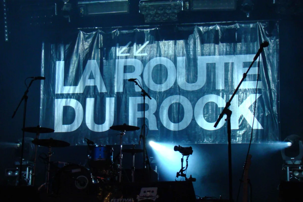 Cédric · 15/08/2009 · 3264 x 2176 Cette photographie montre une scène de concert avec pour sujet principal un écran affichant le texte **"LA ROUTE DU ROCK"** en lettres lumineuses. La composition met en avant des instruments de musique (micro, amplis) et des projecteurs. Le cadrage est serré sur le devant de la scène, avec une légère profondeur de champ.Les lumières sont tamisées et bleutées, créant une ambiance immersive et dynamique. Les couleurs dominantes sont les tons froids (bleu, noir), renforçant une atmosphère électrique et rock. Le style évoque un concert intimiste ou un événement musical organisé.