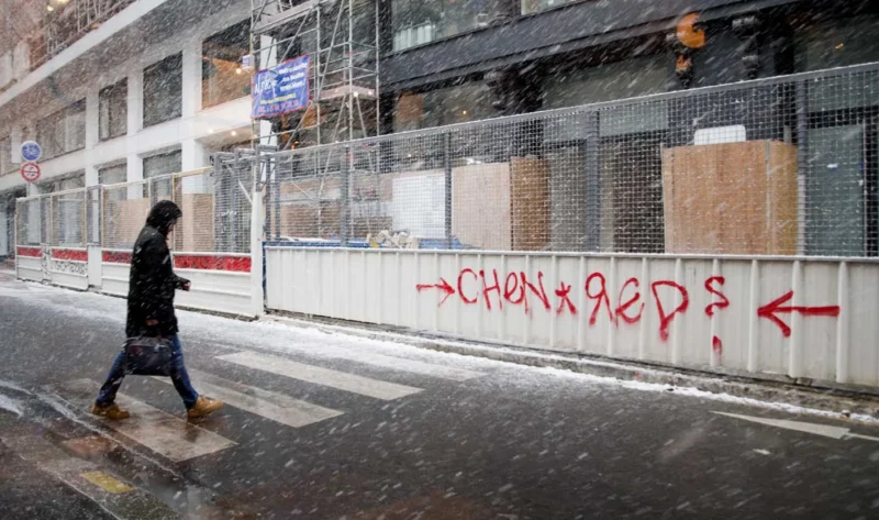 Franck T. · 05/01/2026 · 1400 x 827 Cette photographie montre une personne marchant sous la pluie devant un mur de chantier partiellement recouvert de graffitis. Le sujet principal est cette silhouette floue, suggérant mouvement et solitude.La composition est simple, avec un cadrage serré sur le mur et la personne en arrière-plan. Les couleurs dominantes sont les tons grisâtres de la pluie et du béton, adoucis par les éclats de rouge et de blanc du graffiti. La lumière, diffuse et froide, accentue l’ambiance mélancolique et urbaine. Le style est réaliste et documentaire, renforçant une atmosphère de désolation et de résilience.