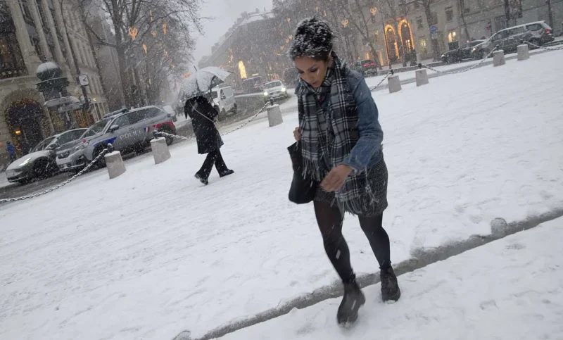 Franck T. · 06/01/2026 · 1400 x 845 Cette photographie capture une femme comme sujet principal, marchant sous la neige en ville. Elle porte un manteau bleu, un bonnet et des gants, et semble observer son téléphone.La composition montre une rue enneigée avec des voitures et des passants en arrière-plan. Le cadrage est serré sur la femme, mais inclut aussi des éléments urbains.La lumière est naturelle, tamisée par la neige et le temps couvert, créant une ambiance froide et grise. Les couleurs dominantes sont les tons froids : blanc de la neige, gris du ciel et des vêtements, avec des touches de bleu et noir.Le style est documentaire et réaliste, évoquant une ambiance hivernale et urbaine.