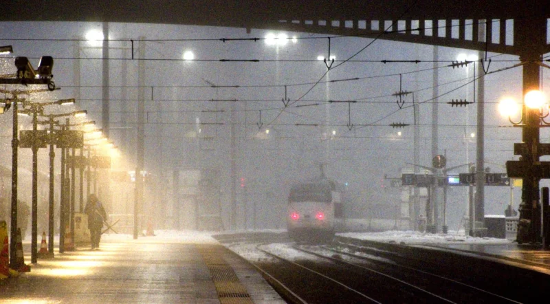 Franck T. · 07/01/2026 · 1400 x 773 Cette photographie capture une gare sous la neige ou le brouillard. Le **sujet principal** est un train en mouvement, éclairé par ses phares arrière rouges. La **composition** montre une voie ferrée centrale, encadrée par des poteaux électriques et des abribus déserts.Le **cadrage** est serré, mettant en valeur la perspective des rails convergeant vers l’horizon. La **lumière**, artificielle et tamisée, crée une ambiance nocturne ou crépusculaire. Les **couleurs dominantes** sont des tons froids : gris, blanc (neige/brouillard) et rouge (feux du train).Le **style** est réaliste et documentaire, évoquant une **ambiance** mélancolique et solitaire.