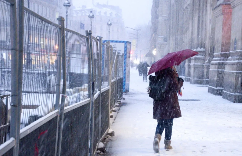 Franck T. · 07/01/2026 · 1400 x 904 Cette photographie montre une personne comme sujet principal, marchant sous une pluie ou neige fine avec un parapluie rouge. La composition met en valeur une perspective longitudinale, encadrée par des barrières métalliques et des bâtiments en arrière-plan.Le cadrage est serré, centré sur la silhouette. La lumière est faible et diffuse, créant une ambiance grise et froide. Les couleurs dominantes sont les tons neutres (gris, blanc) rehaussés par le rouge vif du parapluie.Le style est réaliste et documentaire, évoquant une atmosphère urbaine mélancolique et solitaire.