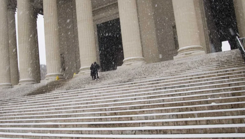 Franck T. · 06/01/2026 · 1400 x 793 Cette photographie capture un **sujet principal** discret : deux silhouettes marchant sur un large escalier enneigé. La **composition** repose sur les lignes architecturales des colonnes et des marches, créant une perspective profonde. Le **cadrage** est serré sur les marches, avec un arrière-plan flou.La **lumière** est douce et diffuse, typique d’un jour nuageux ou d’une neige tombante. Les **couleurs dominantes** sont les tons froids : gris bleuté de la neige, blanc des colonnes et noir des silhouettes.Le **style** est sobre et réaliste, évoquant une **ambiance** calme et mélancolique, presque intemporelle.