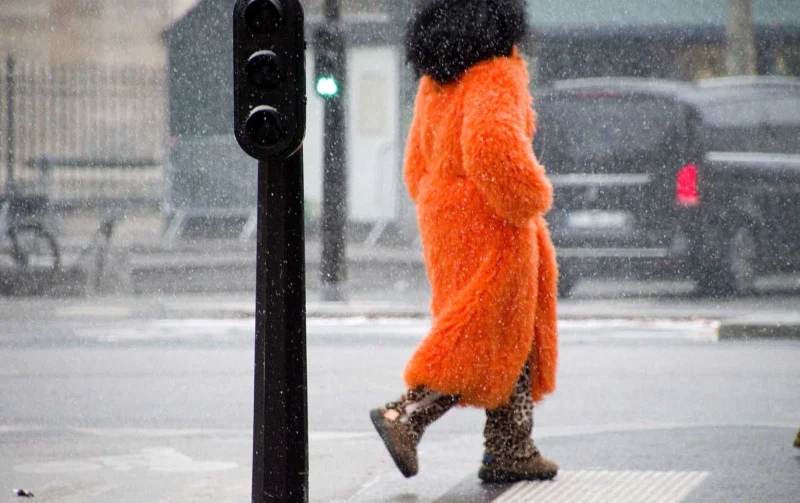 Franck T. · 05/01/2026 · 1400 x 880 Cette photographie met en scène une femme en mouvement, vêtue d'une longue robe orange vif, son visage partiellement caché par une écharpe. La composition est dynamique, centrée sur le sujet qui traverse un carrefour sous la pluie.Le cadrage serré isole la personne, avec un arrière-plan flou comprenant des bâtiments et des véhicules. La lumière est tamisée, typique d’un jour gris et pluvieux, créant une ambiance froide et humide. Les couleurs dominantes sont les tons gris-beige du paysage et l’orange éclatant de la robe, qui contraste fortement. Le style est réaliste et spontané, évoquant une atmosphère urbaine et mélancolique.