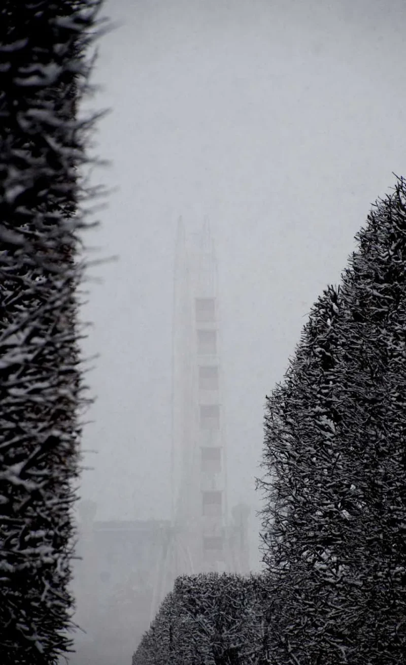 Franck T. · 06/01/2026 · 855 x 1400 Cette photographie met en scène une tour floue, probablement un monument emblématique, partiellement visible à travers un brouillard dense. La composition est centrée sur cette silhouette, encadrée par des haies taillées en forme de colonnes.Le cadrage est serré, accentuant l'effet mystérieux et immersif. La lumière est tamisée, créant une ambiance voilée et onirique. Les couleurs dominantes sont les gris et blancs, renforçant l'atmosphère froide et brumeuse. Le style est épuré et minimaliste, évoquant une touche poétique et contemplative.