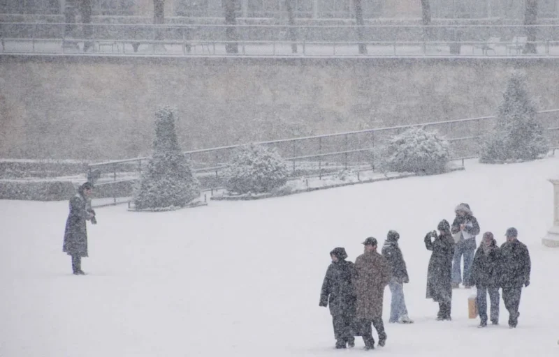Franck T. · 06/01/2026 · 1400 x 896 Cette photographie capture un groupe de personnes marchant dans la neige sous une ambiance hivernale. Le sujet principal est les promeneurs, vêtus chaudement, en premier plan.La composition montre un espace ouvert avec un monument enneigé en arrière-plan. Le cadrage est large, incluant une perspective profonde. La lumière est diffuse, typique d’un jour nuageux, adoucissant les contours.Les couleurs dominantes sont les tons blancs et gris de la neige, avec des touches sombres des vêtements. Le style est réaliste et documentaire, évoquant une ambiance calme et froide, presque mélancolique.