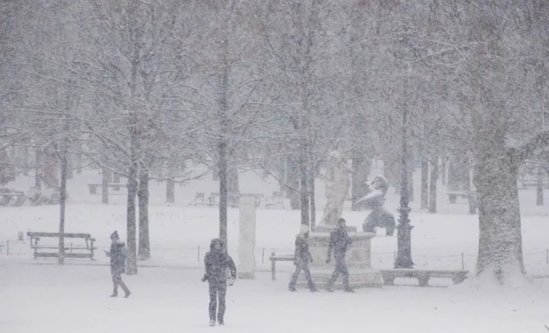 Franck T. · 06/01/2026 · 1400 x 848 Cette photographie capture une scène hivernale en ville. Le **sujet principal** semble être un groupe de personnes marchant sous la neige.La **composition** est équilibrée, avec des arbres dénudés en arrière-plan et des statues ou bancs au centre. Le **cadrage** est large, incluant un espace urbain.La **lumière** est diffuse, typique d’un jour nuageux ou neigeux, adoucissant les contours. Les **couleurs dominantes** sont les tons froids : blanc de la neige, gris du ciel et des arbres, avec des touches sombres sur les vêtements.Le **style** est réaliste et documentaire. L’**ambiance** est calme et mélancolique, évoquant la sérénité d’un hiver urbain.