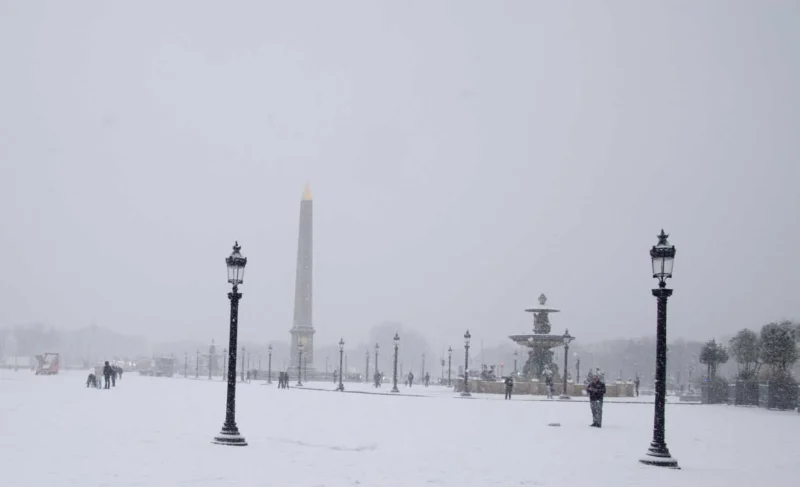 Franck T. · 06/01/2026 · 1400 x 852 Cette photographie montre un paysage hivernal enneigé. Le **sujet principal** est un **monument obélisque** au centre, entouré de lampadaires et d’un petit kiosque à musique.La **composition** est symétrique, avec une perspective axiale menant vers l’obélisque. Le **cadrage** est large, capturant une étendue plane et dégagée. La **lumière** est douce et diffuse, typique d’un jour couvert ou brumeux. Les **couleurs dominantes** sont les tons blancs et gris, renforçant l’ambiance froide et monochrome.Le **style** est réaliste et sobre, évoquant une **ambiance calme et mélancolique**, presque onirique.