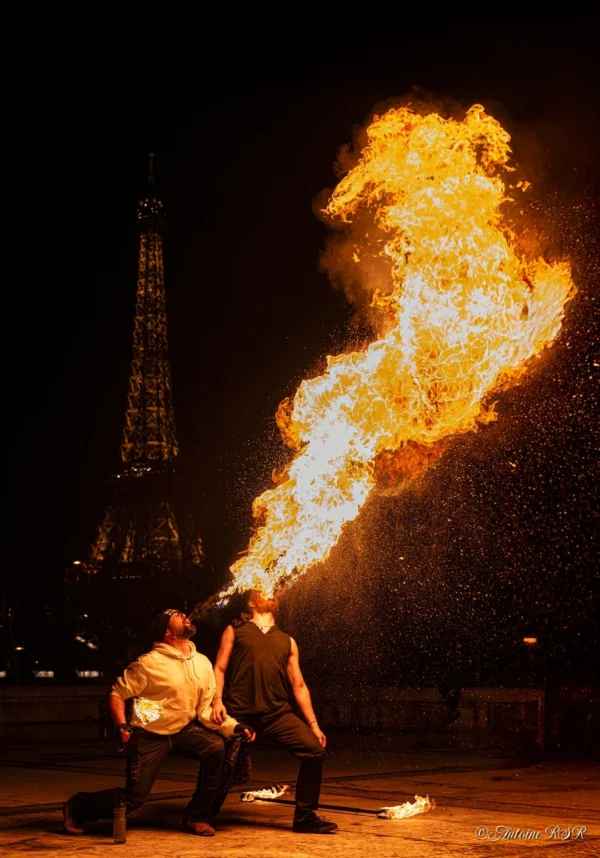 Antoine · 1080 x 1544 Cette photographie capture deux personnes manipulant une torche à feu produisant une flamme géante et courbée. Le sujet principal est cette flamme spectaculaire, en forme de spirale lumineuse.La composition est dynamique, avec un cadrage serré sur les artistes et la flamme, tandis qu'en arrière-plan, on distingue une silhouette de tour Eiffel éclairée. Les couleurs dominantes sont les teintes chaudes et orangées de la flamme, contrastant avec les tons sombres de la nuit. L'ambiance est intense, presque mystique, évoquant un spectacle de feu traditionnel. Le style est réaliste et captivant, mettant en valeur la maîtrise technique des artistes.