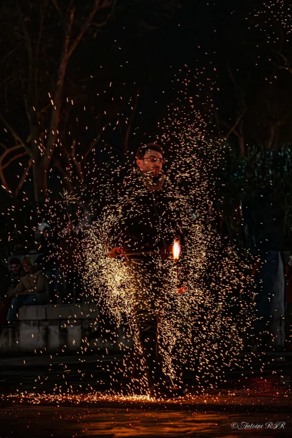 Antoine · 1080 x 1620 Cette photographie capture un individu en mouvement, entouré d’un nuage de particules lumineuses, probablement des étincelles ou des paillettes projetées. Le sujet principal, légèrement flou, est au centre, créant une impression de dynamisme.La composition joue sur le contraste entre le premier plan lumineux et l’arrière-plan sombre. Le cadrage serré isole le sujet, renforçant son impact visuel. Les couleurs dominantes sont les tons chauds (oranges, jaunes) des étincelles, contrastant avec le noir de la nuit.Le style est artistique et dramatique, évoquant une ambiance mystique et festive, presque onirique.