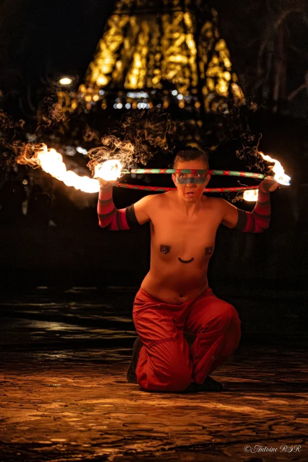 Antoine · 1080 x 1620 Cette photographie met en scène un artiste de feu comme sujet principal, assis en tailleur et manipulant des torches enflammées en cercle autour de lui. La composition est centrée sur ce performer, avec un cadrage serré qui isole son corps.Les flammes et les lumières artificielles créent une ambiance dramatique et mystique. Les couleurs dominantes sont les teintes chaudes des flammes (oranges, jaunes) et le rouge vif des vêtements de l'artiste. Le style est dynamique et captivant, évoquant une scène de spectacle nocturne, presque rituelle.
