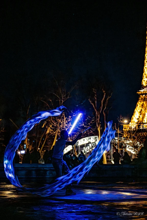 Antoine · 1080 x 1620 Cette photographie capture un **artiste de lumière** en train de dessiner une boucle lumineuse avec une baguette lumineuse. Le **sujet principal** est cette traînée bleue dynamique, contrastant avec l'obscurité.La **composition** met en valeur le mouvement circulaire, encadré par des arbres et l'Eiffel Tower en arrière-plan. Le **cadrage** est serré sur l'action, avec une profondeur de champ réduite.Les **couleurs dominantes** sont le bleu électrique et le noir, créant une **ambiance nocturne mystérieuse et énergique**. Le style relève du **street art photographié**, mêlant créativité et technique.