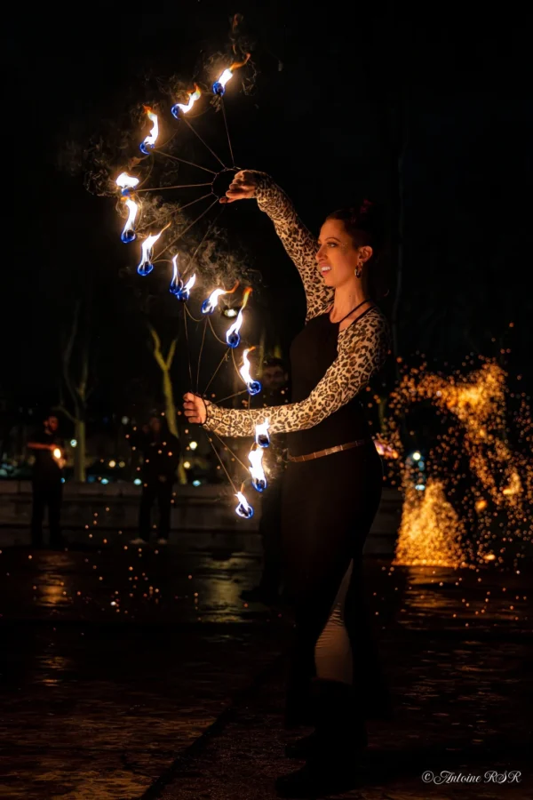 Antoine · 1080 x 1620 Cette photographie met en scène une personne en mouvement, manipulant des torches enflammées. Le sujet principal est dynamique, au centre du cadre, avec un cadrage serré qui isole son action.La lumière est dramatique, avec des éclats vifs des torches contrastant sur un fond sombre. Les couleurs dominantes sont les teintes chaudes des flammes (jaunes, oranges) sur un arrière-plan noir et bleu nuit.Le style est artistique et expressif, évoquant une ambiance mystique et énergique, presque rituelle. L'atmosphère est intense et captivante.