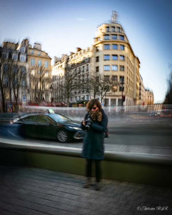 Antoine · 03/01/2026 · 1584 x 1980 Cette photographie met en scène une femme comme sujet principal, vêtue d’une longue veste bleue, absorbée par son téléphone. Le cadrage est serré sur elle, avec un arrière-plan flou de bâtiments urbains et une voiture en mouvement, créant un effet de vitesse.La lumière est douce, probablement naturelle, avec une ambiance crépusculaire ou matinale. Les couleurs dominantes sont les tons froids (bleu, gris) et quelques touches chaudes (briques, éclairage urbain).Le style est artistique, dynamique grâce au flou d’arrière-plan, évoquant une ambiance urbaine moderne et contemplative.