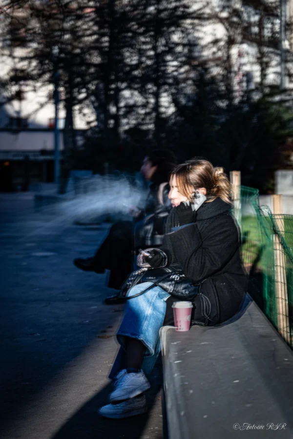 Antoine · 03/01/2026 · 1320 x 1980 Cette photographie met en scène une personne assise seule sur un muret, le visage partiellement caché par ses cheveux et une veste sombre. Le sujet principal semble absorbé, peut-être en train de fumer.La composition est simple et épurée, avec un cadrage serré qui isole le personnage. L’arrière-plan, flou et légèrement surexposé, met en valeur les arbres et un ciel clair.La lumière est douce et naturelle, créant des contrastes subtils. Les couleurs dominantes sont les tons froids (bleus, gris) et les teintes sombres de la veste.Le style est réaliste et légèrement mélancolique, évoquant une ambiance calme et introspective.