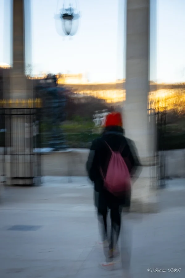 Antoine · 03/01/2026 · 1320 x 1980 Cette photographie capture une personne en mouvement, floue, suggérant une marche rapide. Le sujet principal est un individu avec un sac rouge à dos et un bonnet, partiellement visible sous une colonnade.La composition repose sur des lignes architecturales verticales, encadrant le sujet. Le cadrage est serré, mettant l'accent sur la silhouette et l'arrière-plan flou.Les couleurs dominantes sont les tons froids (gris, bleu) avec des éclats chauds (orange) au loin. La lumière douce, probablement crépusculaire, crée une ambiance paisible et mélancolique. Le style est épuré, presque abstrait, avec un effet dynamique dû au flou de mouvement.