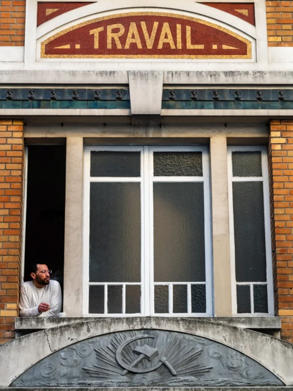 Daniel · 2225 x 2968 Cette photographie montre un homme assis sur un balcon en pierre, regardant vers la gauche. Le sujet principal est donc cet individu, dont le visage est partiellement visible.La composition met en valeur le balcon avec ses grandes fenêtres et la façade en briques. Le cadrage est serré, centré sur le balcon et l’homme. La lumière semble naturelle, avec des ombres douces.Les couleurs dominantes sont les tons beiges des briques, les gris des pierres et les blancs des fenêtres. Le style est réaliste et documentaire, évoquant une ambiance calme et introspective, typique d’un moment de pause ou de réflexion.